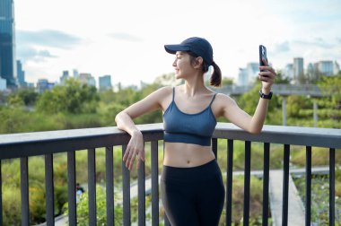 Aktif kıyafetli genç bir kadın şehirde güneşli bir günün tadını çıkarırken akıllı telefonunu kullanıyor..