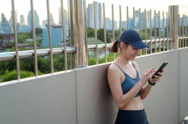 Aktif kıyafetli genç bir kadın şehirde güneşli bir günün tadını çıkarırken akıllı telefonunu kullanıyor..