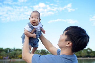 Bir baba neşeli bir şekilde gülümseyen bebeğini açık bir günde parkın tepesinde kaldırıyor..