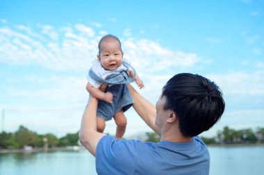 Bir baba neşeli bir şekilde gülümseyen bebeğini açık bir günde parkın tepesinde kaldırıyor..