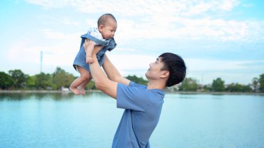 Bir baba neşeli bir şekilde gülümseyen bebeğini açık bir günde parkın tepesinde kaldırıyor..