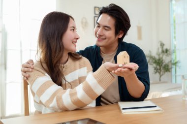Parlak bir yaşam alanında küçük bir ev modelini tutarken çift neşeyle gülümsüyor..