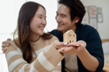 Parlak bir yaşam alanında küçük bir ev modelini tutarken çift neşeyle gülümsüyor..