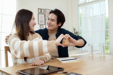 Parlak bir yaşam alanında küçük bir ev modelini tutarken çift neşeyle gülümsüyor..