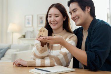 Parlak bir yaşam alanında küçük bir ev modelini tutarken çift neşeyle gülümsüyor..