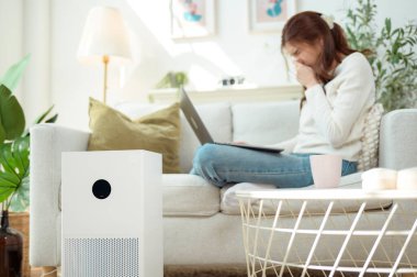 Kanepede oturan bir kadın dizüstü bilgisayarı üzerinde çalışırken hava temizleyicisini çalıştırıyor..