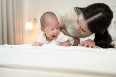 Bir anne, bebeğini, parlak bir odada yatakta emeklemesi için motive ederken gülümsüyor..