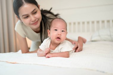 Bir anne, bebeğini, parlak bir odada yatakta emeklemesi için motive ederken gülümsüyor..