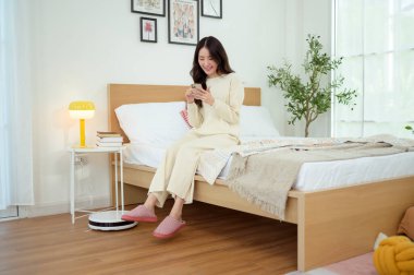 Robotik elektrikli süpürge çalışırken Asyalı genç kadın dinleniyor.