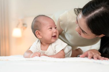 Bir anne, bebeğini, parlak bir odada yatakta emeklemesi için motive ederken gülümsüyor..