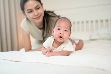 Bir anne, bebeğini, parlak bir odada yatakta emeklemesi için motive ederken gülümsüyor..