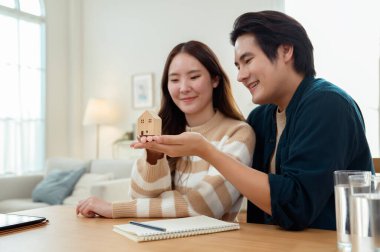 Parlak bir yaşam alanında küçük bir ev modelini tutarken çift neşeyle gülümsüyor..