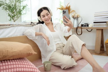 Genç Asyalı kadın yatakta uzanmış akıllı telefon kullanıyor.