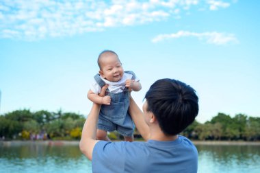 Bir baba neşeli bir şekilde gülümseyen bebeğini açık bir günde parkın tepesinde kaldırıyor..