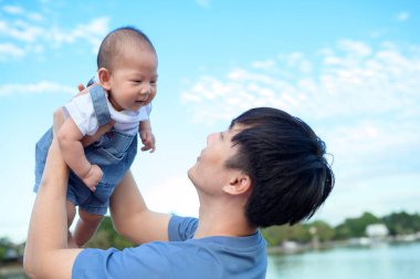 Bir baba neşeli bir şekilde gülümseyen bebeğini açık bir günde parkın tepesinde kaldırıyor..