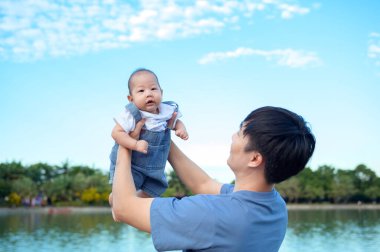 Bir baba neşeli bir şekilde gülümseyen bebeğini açık bir günde parkın tepesinde kaldırıyor..