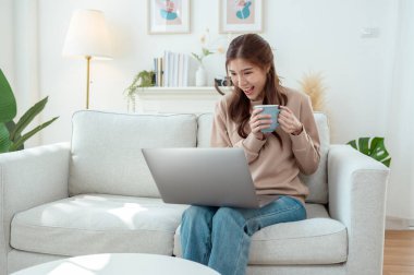 Genç bir kadın dizüstü bilgisayarına odaklanmış, elinde bir fincan kahveyle aydınlık bir oturma odasında.