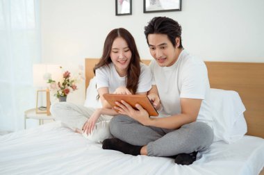 Bir çift, rahatlama anlarında tabletlerinde bir şey izlerken yatakta oturup gülümserler..