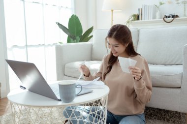 Bir kadın dizüstü bilgisayarı üzerinde çalışırken yerde oturuyor, gülümsüyor ve faturaları düzenliyor..