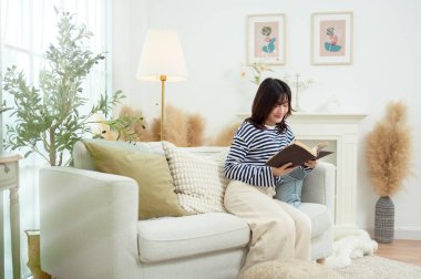 Genç bir kadın kanepede oturuyor, rahat bir ev ortamında bir kitaba dalmış..