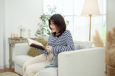 Genç bir kadın kanepede oturuyor, rahat bir ev ortamında bir kitaba dalmış..