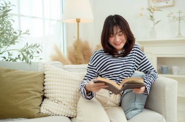 Genç bir kadın kanepede oturuyor, rahat bir ev ortamında bir kitaba dalmış..