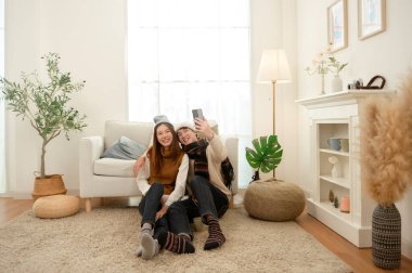 Sıcak bir şekilde giyinmiş iki arkadaş, sıcak bir odada selfie çekerken yere oturup gülümsüyor..