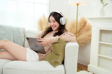 Evdeki oturma odasında tablet kullanan genç Asyalı kadın.