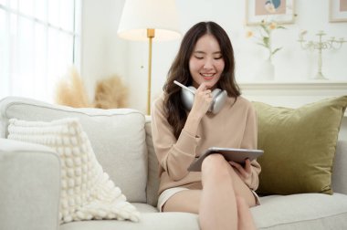 Evdeki oturma odasında tablet kullanan genç Asyalı kadın.