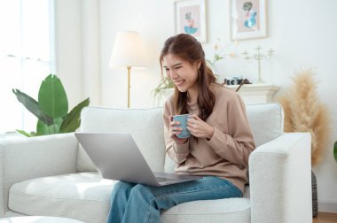 Genç bir kadın dizüstü bilgisayarına odaklanmış, elinde bir fincan kahveyle aydınlık bir oturma odasında.