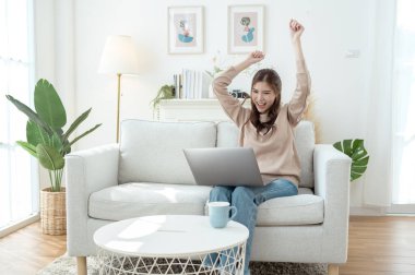 Genç bir kadın dizüstü bilgisayarına odaklanmış, elinde bir fincan kahveyle aydınlık bir oturma odasında.