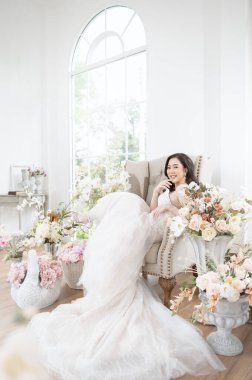 Göz kamaştırıcı bir gelinlik giymiş bir gelin, parlak bir odada çiçeklerin arasında bir anın tadını çıkarır..