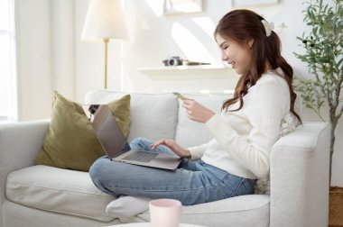 Kahverengi saçlı bir kadın internetten alışveriş yaparken gülümsüyor. Parlak bir oturma odasında rahatça oturuyor..