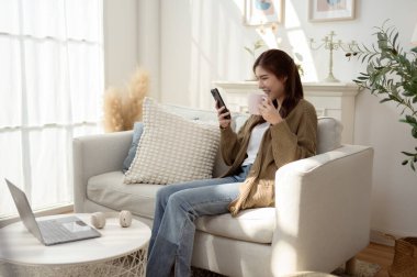 Kadın rahat bir şekilde koltukta oturuyor, parlak ve havadar bir odada akıllı telefonunu kullanıyor..