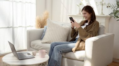 Kadın rahat bir şekilde koltukta oturuyor, parlak ve havadar bir odada akıllı telefonunu kullanıyor..