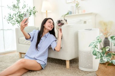 Neşeli bir kadın, rahat bir oturma odasında dinlenirken mutlu köpeğiyle selfie çekmekten hoşlanır..