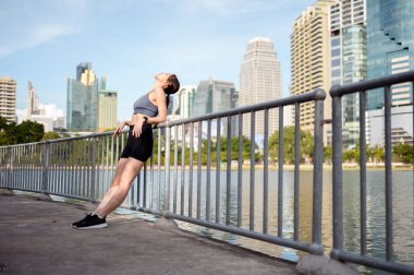 Şehirdeki bir gölün kenarında duran bir kadın, açık hava fitnesinden hoşlanırken sıcak güneşi kucaklıyor..