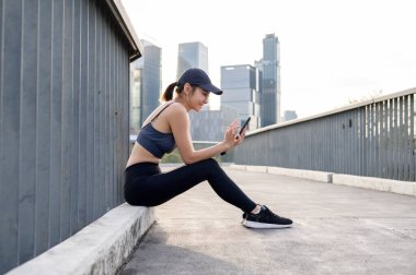 Aktif giyimli bir kadın, modern bir şehir ortamında bir çıkıntıda oturmuş akıllı telefonunu kontrol ediyor..