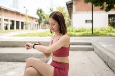 Atletik giyinmiş bir kadın merdivenlerde oturur, parkta gülümseyerek spor saatini kontrol eder..