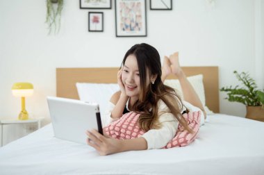 Genç Asyalı kadın yatak odasında tablet bilgisayar kullanmaktan zevk alıyor.