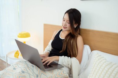 Genç Asyalı kadın yatak odasında dizüstü bilgisayar kullanmaktan zevk alıyor.