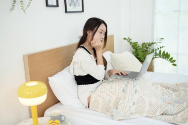 Genç Asyalı kadın yatak odasında dizüstü bilgisayar kullanmaktan zevk alıyor.