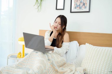Genç Asyalı kadın yatak odasında dizüstü bilgisayar kullanmaktan zevk alıyor.
