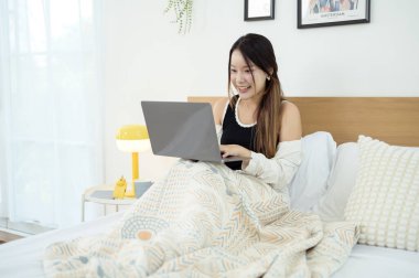 Genç Asyalı kadın yatak odasında dizüstü bilgisayar kullanmaktan zevk alıyor.