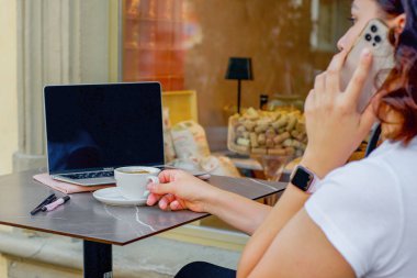 Teras masasında dizüstü bilgisayarla çalışan ve telefonla konuşan genç bir kadın. Yüksek kalite fotoğraf