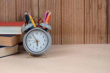 Bir kitap koleksiyonu ve etrafına dağılmış birkaç kalemin yanında duran ahşap bir masanın üzerinde bir çalar saat dinleniyor.