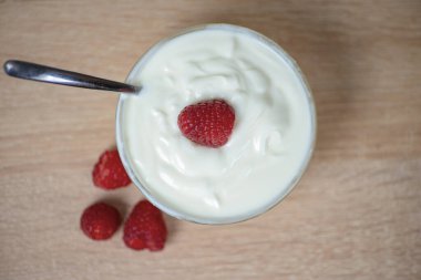 Önünüzde leziz bir kase dolusu kremalı yoğurt var. Üstünde taze ve canlı ahududular var. Yanında da kaşık var.