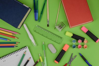 Canlı bir yeşil masa çeşitli okul malzemeleriyle süslenir, renkli öğeler ve öğrenme için gerekli aletler sergilenir