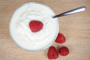 Lezzetli bir kase kremalı yoğurt, yanında da bir kaşık, üzerinde bol bol taze ahududu ve güzel bir şekilde dinleniyor.