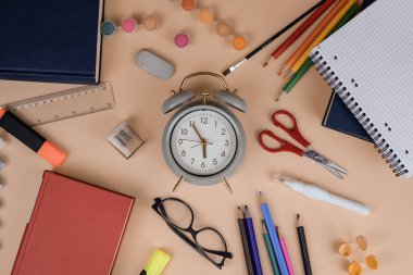Çeşitli okul malzemeleriyle çevrili bir masaya bir çalar saat yerleştirilir ve rahat bir çalışma ortamı oluşturulur.